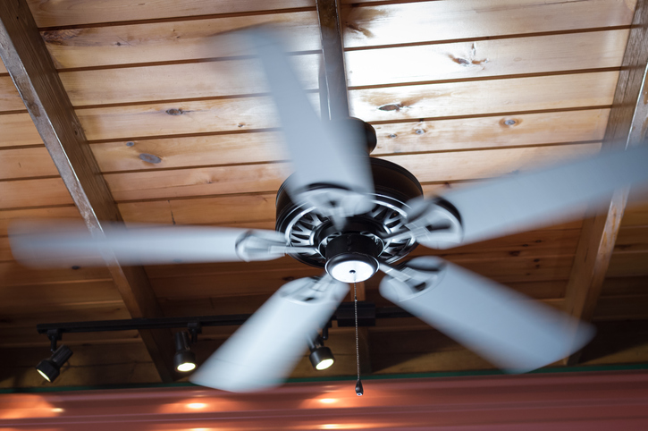 A spinning ceiling fan hangs off of a wooden board ceiling. The fan is turned on and the image exhibits motion blur. The fan is a metallic gray finish.